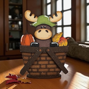 May include: A wooden moose figurine wearing a green hat, peeking over a brown basket. The basket contains a pumpkin and fall leaves. The moose has large antlers and a smiling face. The basket has a handle and is decorated with brown speckles.