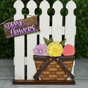 May include: A wooden spring flower sign with a white picket fence and a basket of flowers. The sign reads "Spring Flowers" in purple lettering.