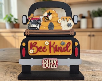 以下が含まれることがあります： 赤い文字で「Bee Kind」と書かれた黄色の荷台が付いた黒い木製のトラックの装飾品。トラックの荷台には、蜂の巣、蜂蜜の瓶、ハニカムが入っています。「Buzz」という言葉がナンバープレートに書かれています。トラックはデイジーと蜂で飾られています。
