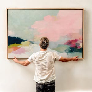 Op de afbeelding: Groot abstract schilderij met een licht houten frame. Het kunstwerk heeft een zacht kleurenpalet van roze, blauw, groen en gele tinten, waardoor een dromerige, wolkenachtige compositie ontstaat. De schilderij wordt door een persoon vastgehouden.