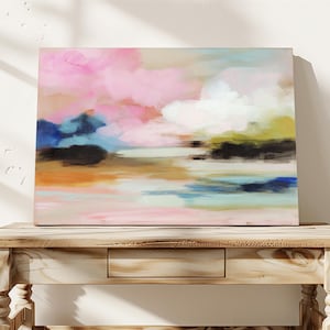 May include: Abstract landscape painting with a palette of pink, blue, and orange hues. The artwork features soft brushstrokes creating a dreamy, cloud-like effect. The canvas is displayed on a light-colored wooden table, adding a touch of rustic charm.