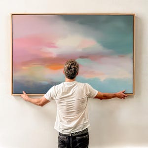 May include: Abstract landscape painting with a wooden frame. The artwork features a blend of colors, including pink, orange, blue, and white, creating a soft, cloud-like effect. The painting is held by a person.