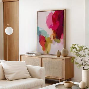 May include: Abstract art print in a wooden frame, showcasing bold pink, red, and yellow paint strokes on white. The artwork is displayed above a wooden cabinet with woven cane doors, alongside neutral-coloured vases.