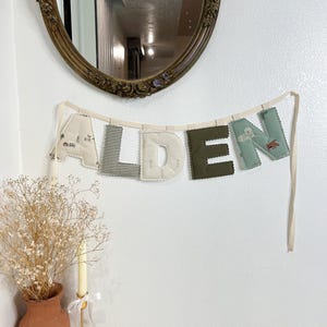 May include: A decorative banner spelling "ALDEN" in fabric letters. The letters are in various colors and patterns, including beige with a car print, black and white stripes, white, olive green, and teal. The banner is suspended by a cream-colored ribbon.