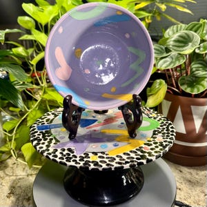 May include: A purple ceramic bowl with pastel-colored abstract shapes, resting on a decorative stand. The stand features a black and white spotted border and a colorful geometric design. The bowl and stand are displayed against a backdrop of green plants.