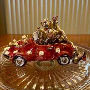 Puede incluir: Figura decorativa de cerámica de un coche descapotable rojo con tapacubos en forma de corazón. El coche está lleno de varias figuras de osos y adornado con pequeñas flores blancas. La figura está colocada sobre un plato de cristal transparente.