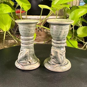 May include: A pair of sage green ceramic candle holders featuring a sculpted frog figure at the base of each piece. The holders have a cylindrical body with three horizontal ridges below the flared top rim and a smooth, glossy glaze finish.