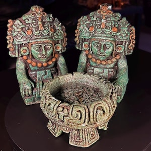 May include: Two identical stone-like figurines featuring detailed headdresses and necklaces, positioned behind a circular, footed bowl with intricate carved geometric patterns. The objects have a weathered, textured green and brown finish.