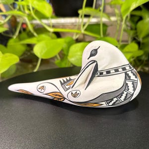 May include: A white ceramic bird figurine featuring black and orange painted geometric patterns and leaf motifs. The bird is depicted in a resting position with its head tucked, showcasing intricate line work and a smooth, matte finish.
