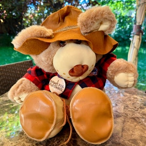May include: A brown teddy bear wearing a brown hat, a red and black plaid shirt, and brown boots. The bear has a button on its chest that says "I Paws For Furskins".