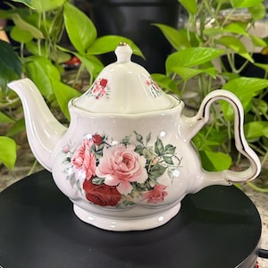 Puede incluir: Una tetera de cerámica color crema con un estampado floral de rosas rosas y rojo oscuro con hojas verdes. La tetera tiene una base festoneada, una tapa a juego con un remate dorado y un asa curva con un borde de color dorado.