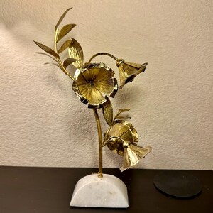 以下が含まれることがあります： 3つの花と葉が付いた、金色の金属製の花の彫刻。彫刻は白い大理石の台座に取り付けられています。花は詳細でテクスチャ加工されたデザインです。装飾的な家のアクセントです。