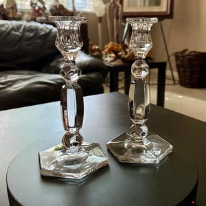 May include: A pair of clear crystal candle holders. Each features a hexagonal base, a faceted stem, and a flared top. The candle holders are placed on a dark surface, reflecting light and creating a sparkling effect. These decorative pieces add elegance to any setting.
