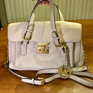 Puede incluir: Un bolso de mano de cuero beige claro con herrajes dorados, que incluyen una cadena y un charm con el logotipo. El bolso tiene un asa superior, un cierre de solapa y una correa para el hombro desmontable. La marca "MICHAEL KORS" es visible.