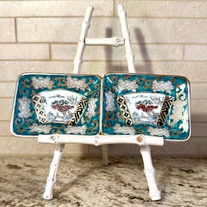 Puede incluir: Dos platos cuadrados decorativos de color turquesa con ribetes dorados y motivos florales. Cada plato presenta un paisaje con un diseño de abanico. Los platos se exhiben en un pequeño caballete de bambú blanco.
