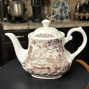 May include: A white ceramic teapot featuring a brown transferware landscape pattern depicting a bridge, trees, and buildings. The lid is topped with a finial and decorated with a floral motif. The teapot has a curved handle and spout.