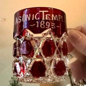 May include: A ruby red glass mug featuring a faceted geometric pattern and a clear glass handle. The upper portion of the mug displays the etched text MASONIC TEMPLE and the year 1893 in a light, contrasting color against the deep red glass surface.
