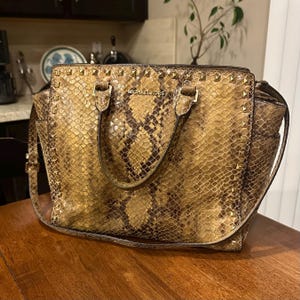 Op de afbeelding: Een Michael Kors handtas met een slangenprint in bruin- en goudtinten. De tas heeft goudkleurige studs langs de boven- en zijkanten, met de merknaam op de voorkant. Hij heeft een handvat aan de bovenkant en een schouderriem.