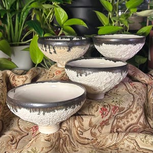 May include: A set of four ceramic bowls with a textured, cratered white exterior and a smooth, cream-colored interior. Each bowl features a dark, metallic-toned rim with a distinctive drip-like glaze pattern extending down the upper edge.