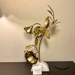 以下が含まれることがあります： 3つの花といくつかの葉が付いた、金色の金属製の花の彫刻。彫刻は白い大理石の台座に取り付けられています。「Papetal」という単語が書かれたタグがベースに付いています。