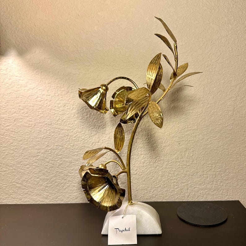 以下が含まれることがあります： 3つの花といくつかの葉が付いた、金色の金属製の花の彫刻。彫刻は白い大理石の台座に取り付けられています。「Papetal」という単語が書かれたタグがベースに付いています。