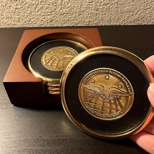 以下が含まれることがあります： 黒色のセンターを持つ金色のコースター4枚セット。各コースターには、翼を広げた金色の鷲、地球儀、そして「Counterterrorism Fellows Program National Defense University」の文字が刻まれています。コースターは木製の箱に入っています。