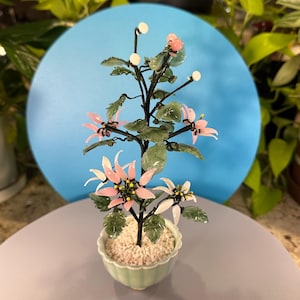 Könnte beinhalten: Ein dekoratives Glasblumenarrangement in einem kleinen, hellgrünen Keramiktopf. Das Arrangement zeigt einen dunkelbraunen Stiel mit rosa und weißen Blüten, grünen Blättern und weißen kugelförmigen Akzenten. Der Hintergrund umfasst einen blauen Kreis und grünes Laub.