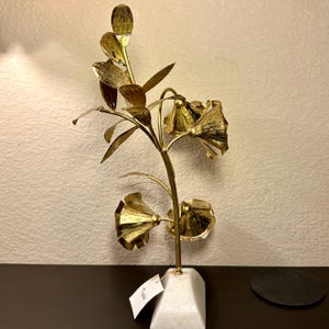 以下が含まれることがあります： 3つの花といくつかの葉が付いた、金色の金属製の花の彫刻。彫刻は、白い幾何学的な大理石の台座に取り付けられています。花は質感があり、細い茎に配置されています。