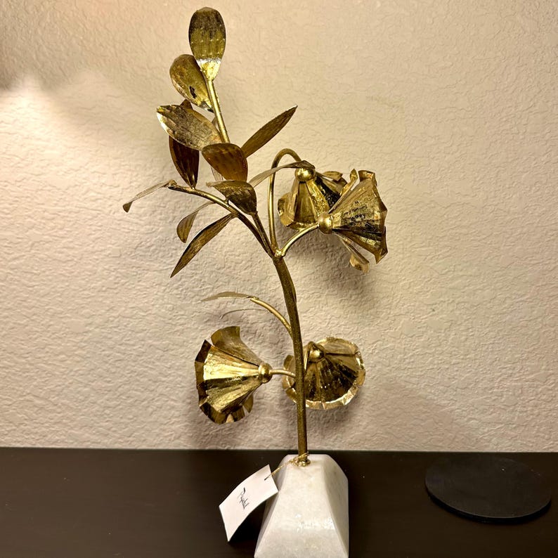以下が含まれることがあります： 3つの花といくつかの葉が付いた、金色の金属製の花の彫刻。彫刻は、白い幾何学的な大理石の台座に取り付けられています。花は質感があり、細い茎に配置されています。