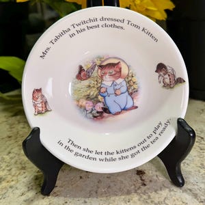 Puede incluir: Plato de cerámica blanca con ilustraciones de gatitos y una mariposa, con texto alrededor del borde. La imagen central muestra un gatito con un traje azul. El plato se exhibe en un soporte negro.