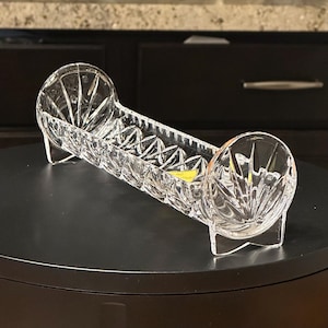 May include: A clear pressed glass celery dish featuring a long rectangular body with a diamond-patterned base and two circular, fan-shaped ends that serve as feet. The glass has a reflective, faceted surface with a small yellow sticker.