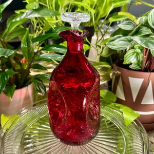 Może przedstawiać: Rubinowoczerwony szklany dekanter z przezroczystym szklanym korkiem. Dekanter ma teksturowaną powierzchnię i unikalny kształt. Korek jest okrągły z rozszerzoną podstawą. Dekanter stoi na przezroczystym szklanym talerzu.