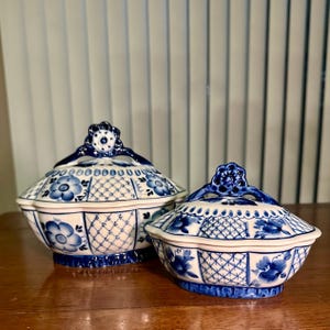 May include: Two white and blue ceramic lidded serving dishes. The dishes have a floral and lattice design with blue accents. The lids have a decorative handle. The dishes are on a wooden surface.