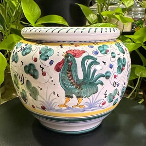 May include: A round ceramic planter with a white base featuring a hand-painted green rooster with a red comb and yellow feet. The design includes teal floral accents, thin black swirls, and yellow and green horizontal bands around the rim and base.