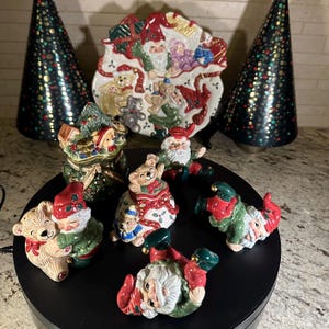 May include: A collection of Christmas-themed figurines and decorations. The set includes Santa Claus and teddy bear figures, a decorative plate, and two cone-shaped Christmas trees. The figurines are painted in festive colors like red, green, and white.