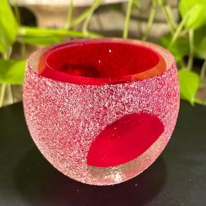 Op de afbeelding: Een bolvormige glazen kaarshouder met een felrode binnenkant en een getextureerd, mat oppervlak aan de buitenkant. De buitenlaag heeft een korrelig, kristallijn uiterlijk in roze en witte tinten, met een glad, helder rood cirkelvormig accent.