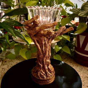May include: A decorative candle holder with a clear, faceted glass bowl supported by a brown, tree-like base. The base has intricate details, and a lit candle is visible inside the bowl. The candle holder is on a black surface.
