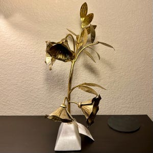 以下が含まれることがあります： 3つのベル型の花といくつかの葉が付いた、金色の金属製の花の彫刻。彫刻は白い幾何学的な台座に取り付けられています。花は質感のある外観をしています。