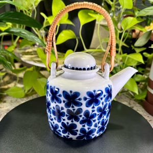 May include: A white ceramic teapot with a blue floral pattern. The teapot has a curved spout, a rounded lid, and a woven handle. The floral design is a repeating pattern of stylized blue flowers.