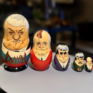 Könnte beinhalten: Ein Set aus fünf hölzernen Matroschka-Puppen mit karikierten Porträts politischer Persönlichkeiten. Die größte Puppe zeigt die russische Flagge und kyrillische Schrift, während die zweite einen roten Anzug mit kyrillischem Text trägt.