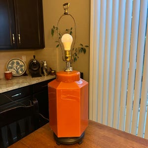 May include: A vibrant orange, geometric table lamp with a gold-coloured neck and a white lightbulb. The lamp is set on a dark brown base and placed on a wooden table. The lamp has a vintage design.