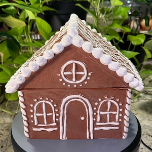 May include: A brown ceramic gingerbread house cookie jar with white icing details. The house features a central arched door, two rectangular windows, a circular window in the gable, and white piped icing along the roofline and edges.