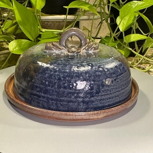 May include: A ceramic butter dish with a dome-shaped lid featuring a deep blue, speckled glaze and a central loop handle. The base is a flat, circular plate with a reddish-brown, earthy rim that contrasts with the glossy blue finish of the cover.