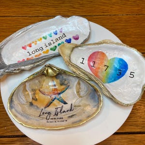Könnte beinhalten: Drei Muschelschalen als Schmuckschalen. Eine hat ein Regenbogenherz mit den Zahlen 11735, eine hat einen Seestern und den Text "Long Island, New York 40.7891° N, 73.1350° W" und eine hat den Text "long island" mit einer Reihe bunter Herzen.