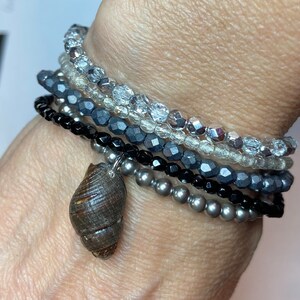 May include: A stack of bracelets with different colored beads, including black, gray, and silver. A small brown seashell charm hangs from the bottom of the stack.