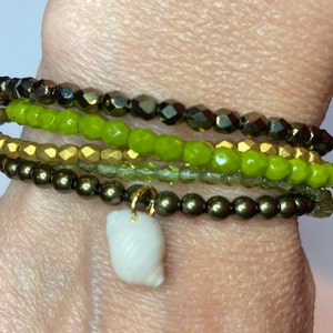 May include: A stack of bracelets with green, gold, and brown beads. The bottom bracelet has a white seashell charm.