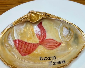 May include: A seashell with a gold rim and a painted red mermaid tail. The shell is about 2 inches wide and has the words "born free" written on it.