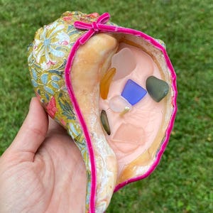 Peut inclure: Une coquille de mer avec un motif floral et une bordure en velours rose, remplie de verre de mer et de pierres.