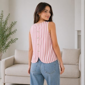 May include: Sleeveless top with vertical stripes in white and pink. The back of the top features a row of six round, light-colored buttons. The top is paired with light blue denim jeans.
