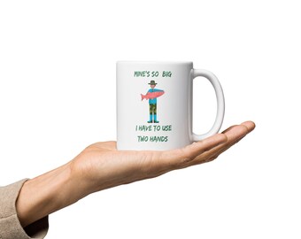 May include: White ceramic mug with a green and brown cartoon illustration of a person holding a pink fish. The text on the mug reads "Mine's so big I have to use two hands."